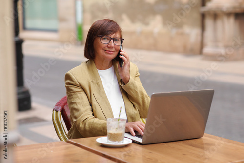 Mature office employee woman talks on smartphone with smile about upcoming business trip while sitting on terrace in cafe with cup of coffee and laptop, happy businesswoman working remotely online.