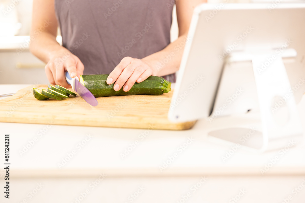 A woman is cutting a cucumber on a cutting board. A tablet is on a stand behind her
