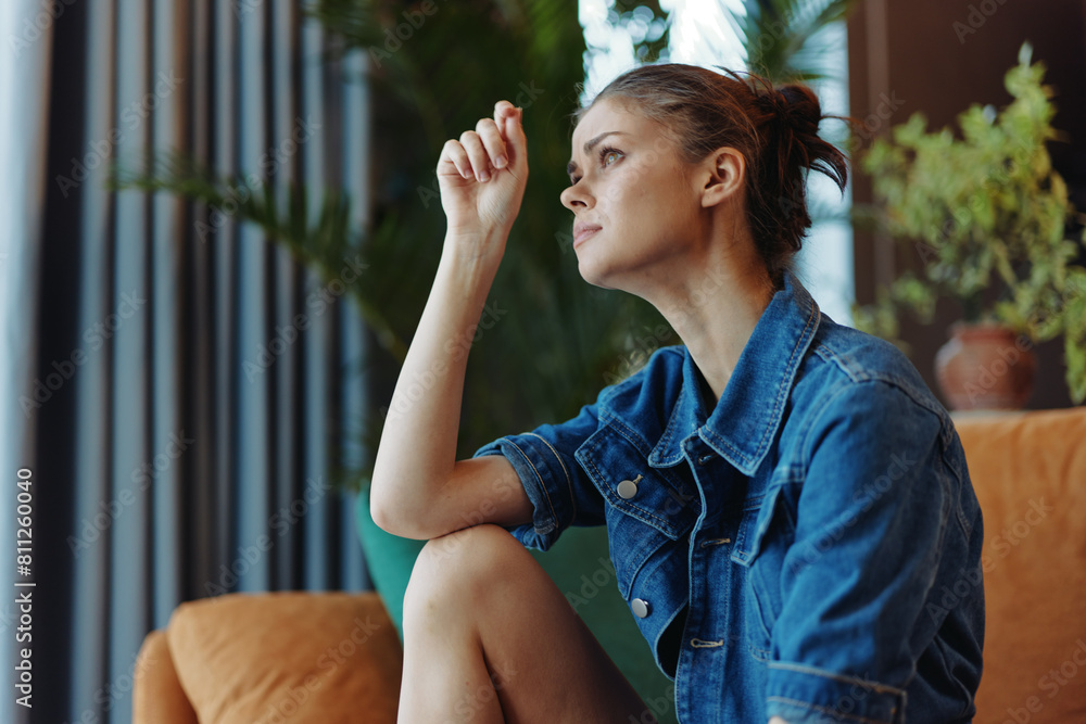 Contemplative woman sitting on couch with hand on face looking out window in thoughtful reflection Contemplative woman sitting on couch with hand on face looking out window in thoughtful reflection