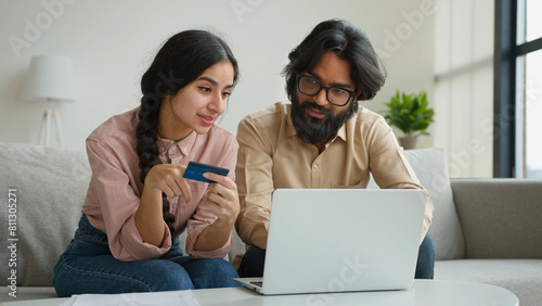 Arabian Indian muslim couple family sit on sofa in living room home office use credit bank card typing code number in laptop banking app pay online multiethnic woman and man shopping internet delivery