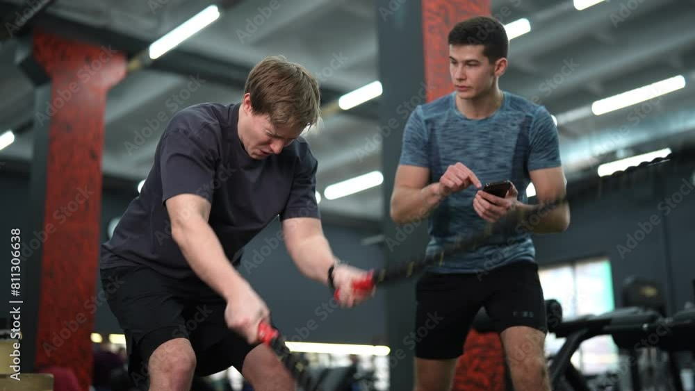 Low-angle view of dedicated beginner sportsman exercising with battle rope while coach encouraging during cross training in health club standing with stopwatch. Shooting in slow motion. Low-angle view of dedicated beginner sportsman exercising with battle rope while coach encouraging during cross training in health club standing with stopwatch. Shooting in slow motion.