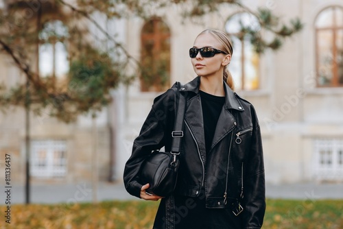 A serious woman with bun hair and glasses on her eyes is wearing a black t-shirt, a black leather jacket and leather pants, holds a black bag on her shoulder, walks in the park © JJ Studio