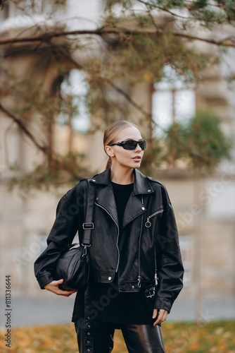 Fashionable woman with black sunglasses on her eyes is looking away from the camera and is wearing a black basic t-shirt, black leather jacket and black leather pants, holding a black bag 