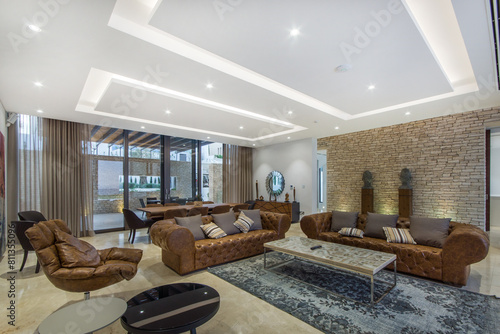 Elegant and contemporary luxury villa living room with leather sofas and modern lighting. A stone accent wall and large windows complement the plush furnishings.