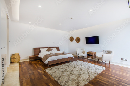 A spacious, bright hotel bedroom with a carpet on the wooden floor, a wide bed, armchairs and a TV. The room is well-lit, with neutral decor and cozy bedding