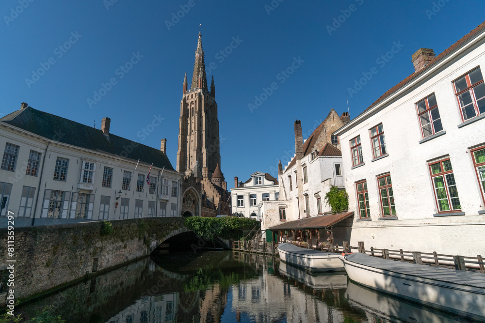 Obraz premium Wharf on the canal since Dijver park with the Our Lady church tower reflected on the canal in the old town of the beautiful city of Brugge in Belgium in a sunny day.