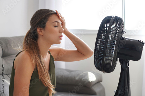 Overheated young woman in front of the working fan suffering from summer heat at home. Girl sitting in front of ventilator in hot weather. Hot summer concept.