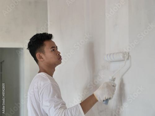 Asian male painter painting new white wall with roller brush, house interior renovation