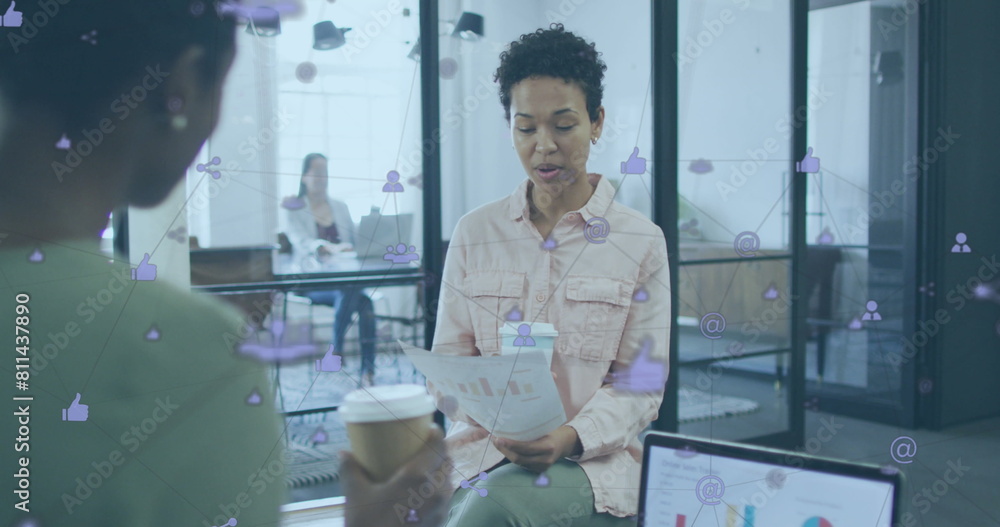 Image of connected icons over diverse female coworkers discussing reports while drinking coffee Image of connected icons over diverse female coworkers discussing reports while drinking coffee