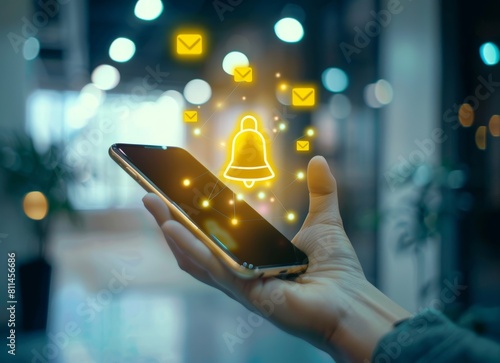 Hand holding a smartphone with a digital glowing yellow bell icon and message icons floating above on a blurred office background, depicting an online communication concept.