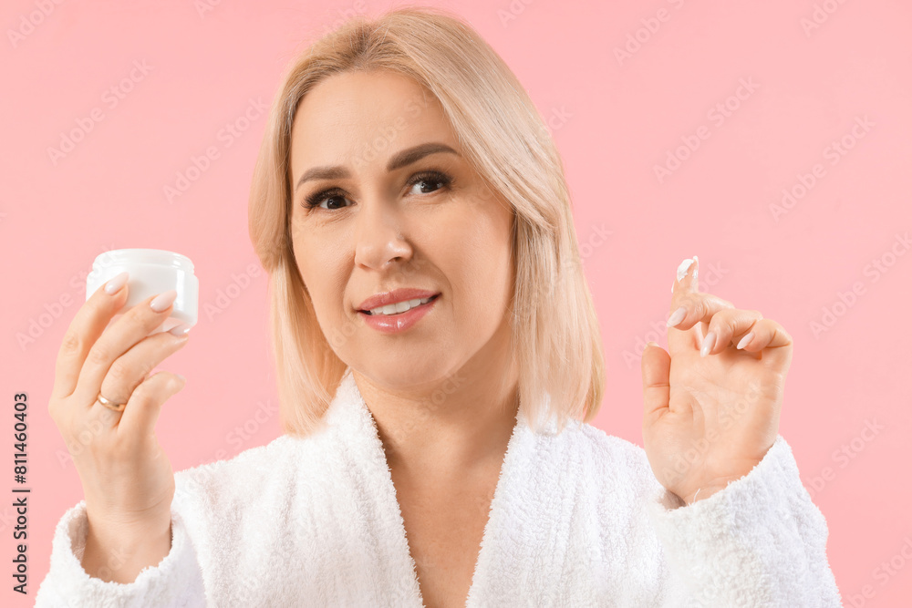 Beautiful mature happy woman in bathrobe with jar of cream on pink background Beautiful mature happy woman in bathrobe with jar of cream on pink background