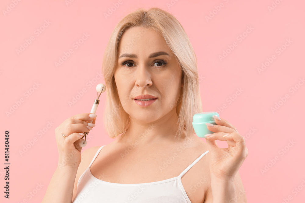 Beautiful mature woman with jar of facial cream and facial massage tool on pink background Beautiful mature woman with jar of facial cream and facial massage tool on pink background