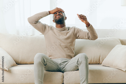 A tired African American man sitting on a couch at home, feeling stressed and frustrated due to a headache He appears sick and overwhelmed, showing signs of physical and mental fatigue A sense of photo