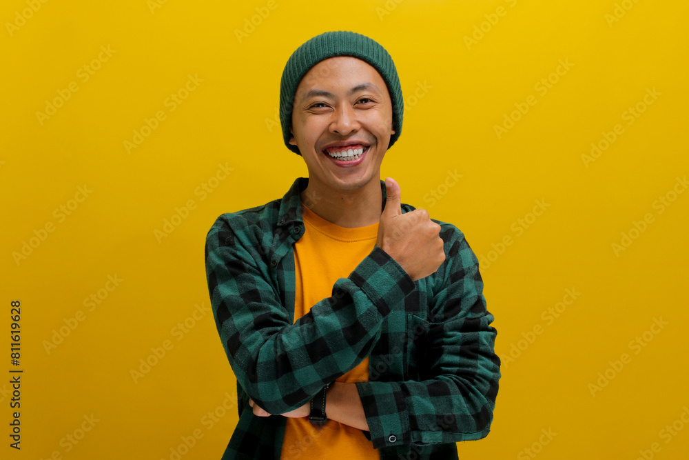 A smiling young Asian man, dressed in casual clothes and wearing a beanie hat, is giving a thumbs up gesture, indicating a positive or good review while standing against a yellow background