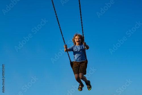 Child swinging on chain swing on city kids playground. Swing ride. Cute child having fun on a swing on summer sky background. Blonde little boy swings at kid playground. Child swinging high.