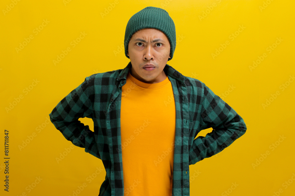 Angry young Asian man, wearing a beanie hat and casual outfit, stands with arms akimbo, looking furious at the camera. Isolated on a yellow background Angry young Asian man, wearing a beanie hat and casual outfit, stands with arms akimbo, looking furious at the camera. Isolated on a yellow background