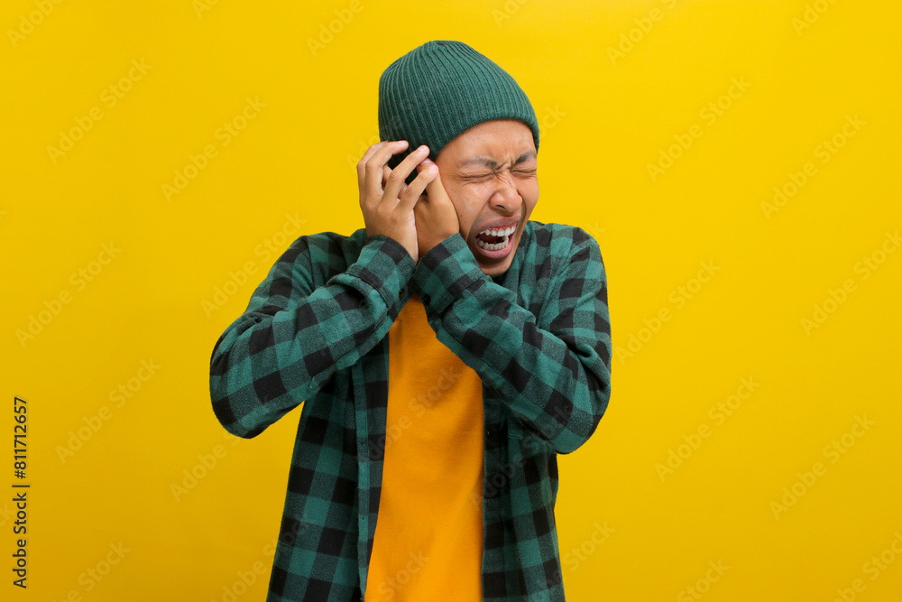 Annoyed young Asian man, wearing a beanie hat and casual shirt, covers his ears and appears to be screaming or shouting, experiencing a painful earache possibly due to otitis exacerbated by loud noise Annoyed young Asian man, wearing a beanie hat and casual shirt, covers his ears and appears to be screaming or shouting, experiencing a painful earache possibly due to otitis exacerbated by loud noise