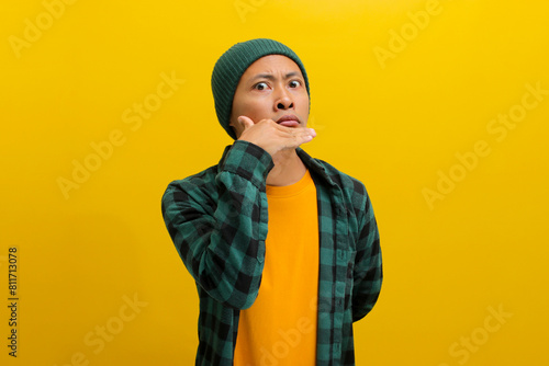 An annoyed young Asian man, dressed in a beanie hat and casual shirt, is making an Italian hand gesture that signifies indifference or disregard, while standing against a yellow background