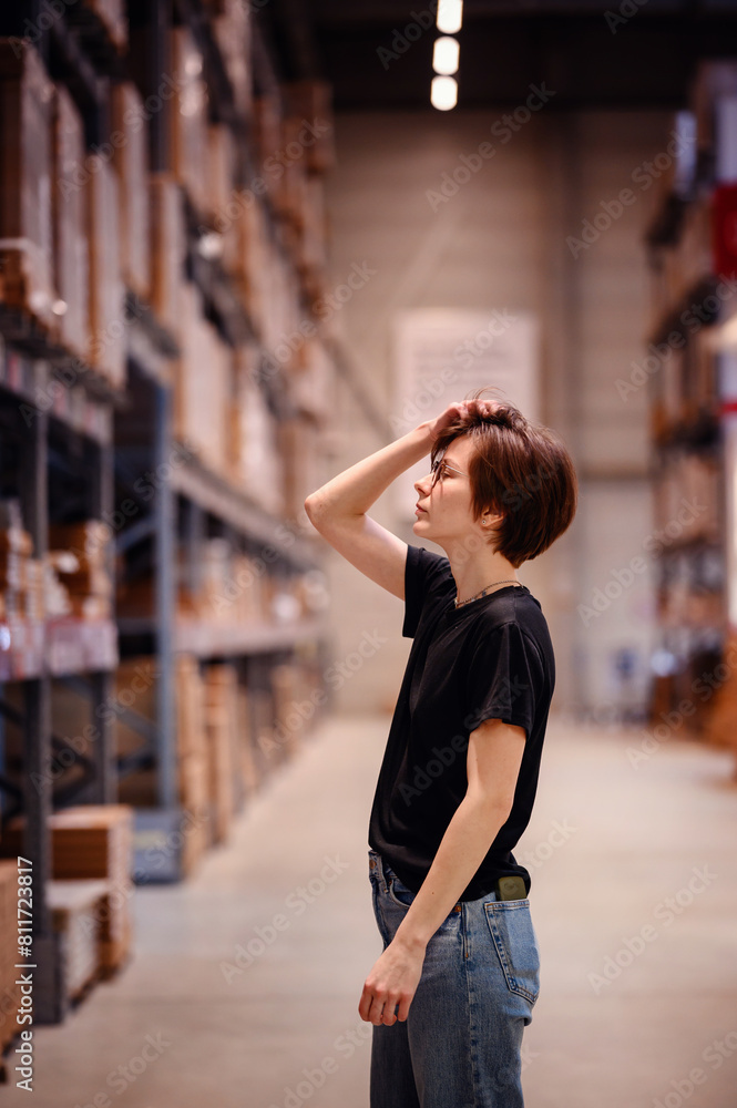 Captured in a spacious warehouse, young woman in casual attire, reaching for an item on a high shelf. 