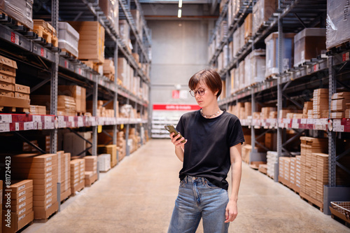 Young woman engaging with her smartphone while standing in a warehouse aisle. Her focused expression and casual style, complete with glasses and modern attire, 