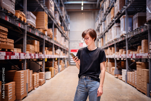 Young woman engaging with her smartphone while standing in a warehouse aisle. Her focused expression and casual style, complete with glasses and modern attire, 