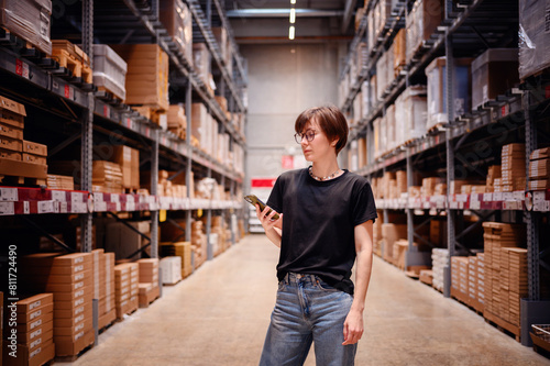 Young woman confidently walking down a warehouse aisle, smartphone in hand, possibly checking product locations or inventory details. Her modern