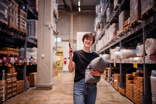 Young woman using her smartphone while shopping in a warehouse, holding a cushion in one arm. Her focused look at the phone screen 