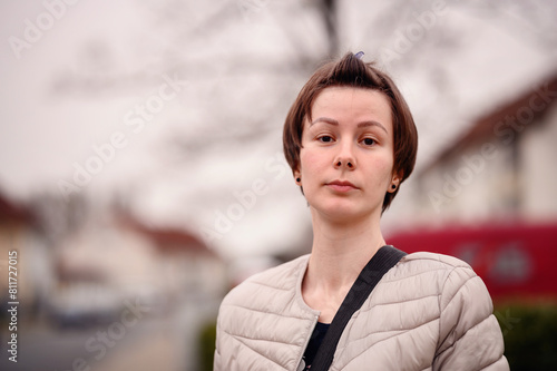 A young woman with short brown hair gazes contemplatively while standing in a suburban neighborhood. She wears a light beige quilted jacket and carries a black shoulder strap photo