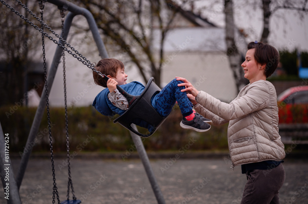 Obraz premium A heartwarming moment of a mother pushing her young son on a swing at a local park. The boy, laughing with glee, reaches out his feet to meet his mother's hands in a playful game. 