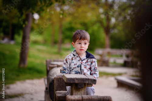 This image beautifully captures a pensive young boy sitting in a wooden structure at a park, surrounded by lush greenery. His contemplative gaze 