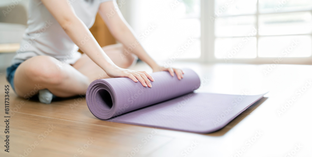 Fototapeta premium Young yoga Woman rolling her lilac mat after a yoga, sitting on wooden floor near a window,