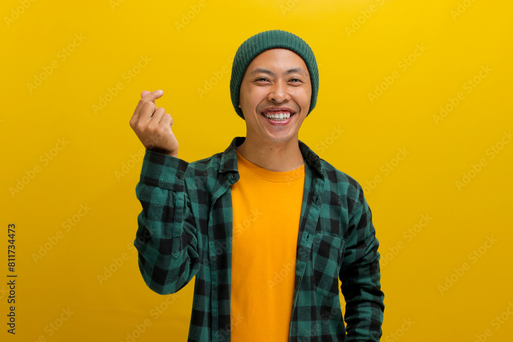 Attractive young Asian man, dressed in beanie hat and casual shirt, exudes happiness as he creates a romantic mini heart shaped gesture with his finger, expressing tender feelings, isolated on yellow Attractive young Asian man, dressed in beanie hat and casual shirt, exudes happiness as he creates a romantic mini heart shaped gesture with his finger, expressing tender feelings, isolated on yellow