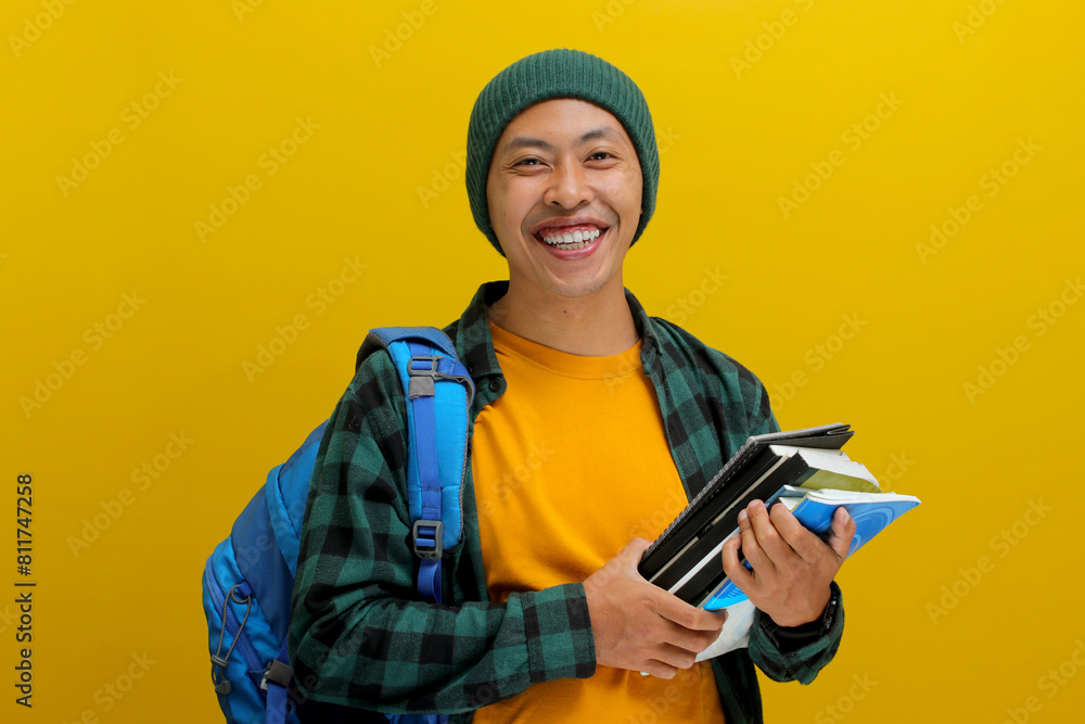 A confident young Asian student, dressed in a beanie hat and casual shirt and carrying a backpack, looks happy while holding a pile of books and standing against a yellow background A confident young Asian student, dressed in a beanie hat and casual shirt and carrying a backpack, looks happy while holding a pile of books and standing against a yellow background