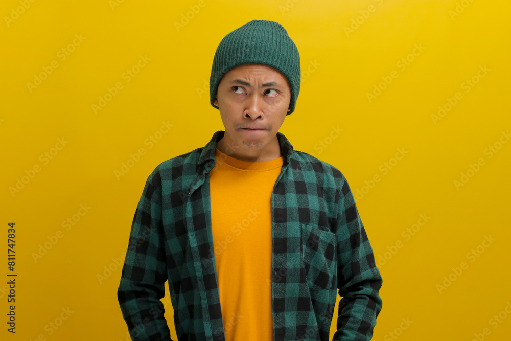 Confused young Asian man is pursing or pinching his lips, displaying doubt and uncertainty while refraining from sharing important information. He appears hesitant, glancing sideways at copy space Confused young Asian man is pursing or pinching his lips, displaying doubt and uncertainty while refraining from sharing important information. He appears hesitant, glancing sideways at copy space