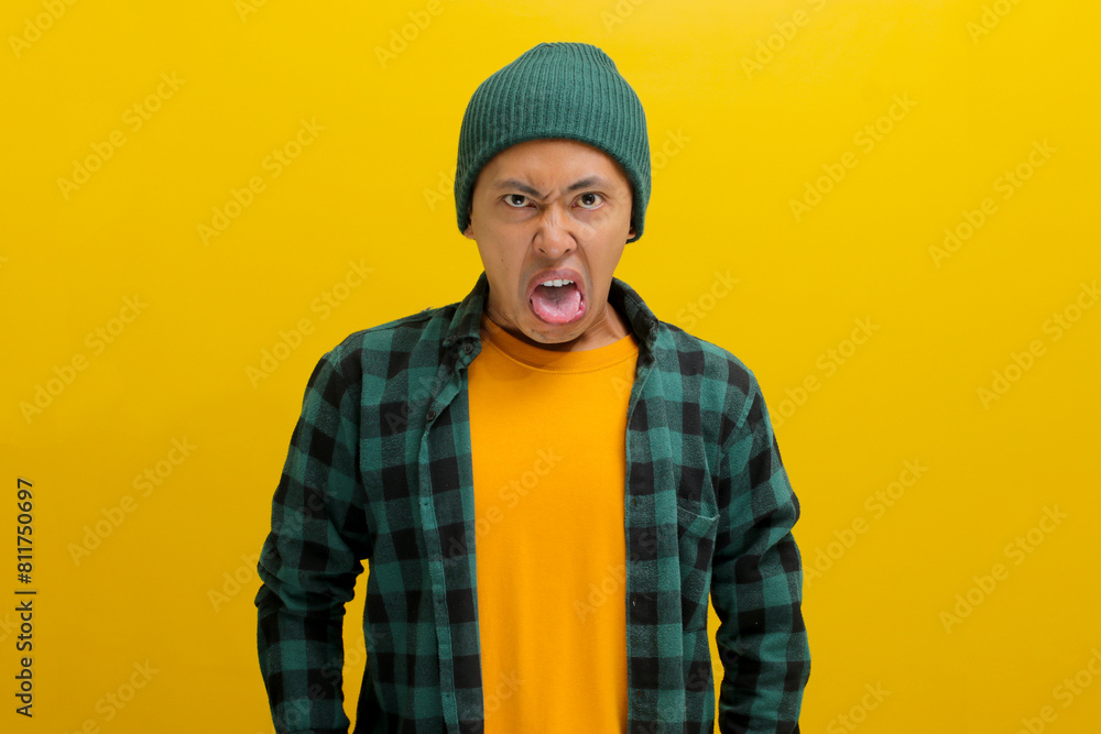 Displeased young Asian man, dressed in a beanie hat and casual shirt, is sticking out his tongue in disgust while standing against a yellow background Displeased young Asian man, dressed in a beanie hat and casual shirt, is sticking out his tongue in disgust while standing against a yellow background