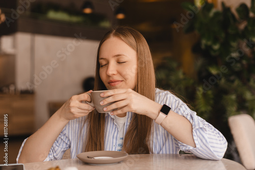 Portrait of gorgeous smiling lady with closed eyes smelling, enjoying of coffee and drinking cappuccino from cup while resting in restaurant. Woman wear stripped shirt.