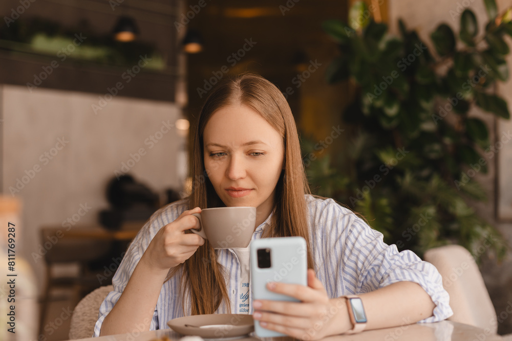 Young beautiful woman holding mobile and looking at smartphone while sitting at cafeteria. Happy girl using mobile phone and look flirting. Woman drinking coffee and smiling, sitting in cafe. Young beautiful woman holding mobile and looking at smartphone while sitting at cafeteria. Happy girl using mobile phone and look flirting. Woman drinking coffee and smiling, sitting in cafe.