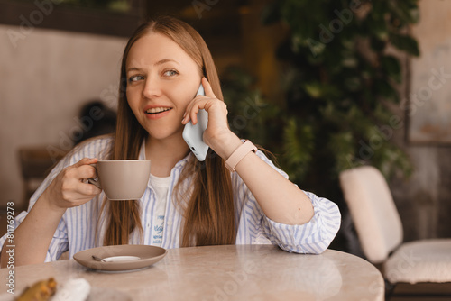 Blonde woman smiling positive conversation talking to her friend using phone hand hold hot drink coffee relax casual positive emotion weekend relax routine at cafe restaurant. Girl have discussion.
