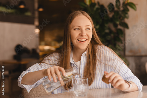 Woman pours water into a glass from a glass bottle. Blonde woman have a good time in cafe, she look happy and smiling. Girl wear stripped shirt.