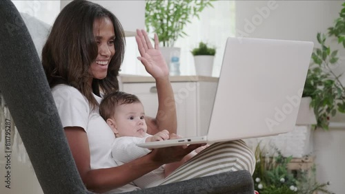 Smiling mixed race woman video conference with friends or relatives holding baby in her arms and waving affably at laptop camera photo