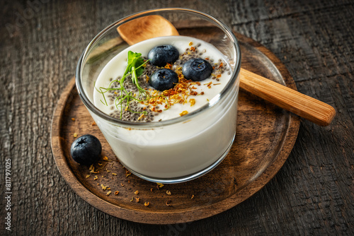 Glass glass with homemade yogurt on a wooden saucer with a wooden spoon on a textured gray wooden background with blueberries, chia seeds, dried osmanthus flowers and pea sprouts. Side and top view.