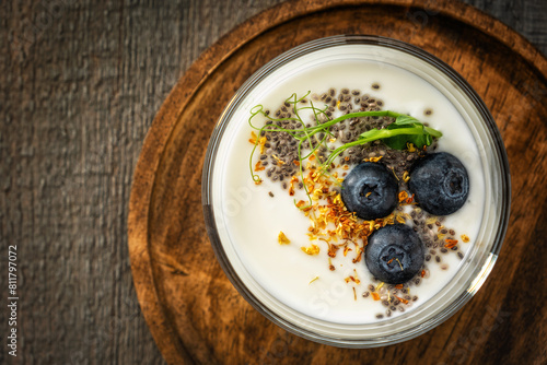 Close-up of homemade yogurt in a glass cup decorated with blueberries, chia seeds, osmanthus flowers and pea sprouts on a round textured wooden plate on a gray wooden background. View from above.