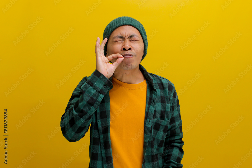Joyful young Asian man, closes his eyes and uses his fingers to make a gourmet or delicious hand gesture, expressing how enjoyable the food is while standing against a yellow background Joyful young Asian man, closes his eyes and uses his fingers to make a gourmet or delicious hand gesture, expressing how enjoyable the food is while standing against a yellow background