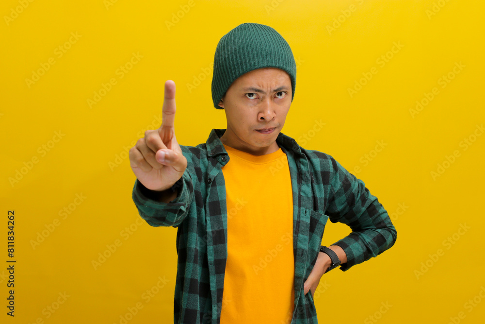 Serious and upset young Asian man, dressed in a beanie hat and casual shirt, is frowning while gesturing a STOP gesture with one finger, expressing disapproval or refusal towards something. Taboo sign Serious and upset young Asian man, dressed in a beanie hat and casual shirt, is frowning while gesturing a STOP gesture with one finger, expressing disapproval or refusal towards something. Taboo sign