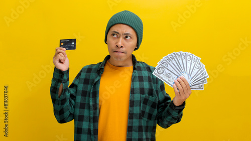 Contemplative Asian man in a beanie and casual clothes examines a credit card and cash, pondering a purchase. Isolated on yellow background. Financial decisions, budgeting, and thoughtful spending