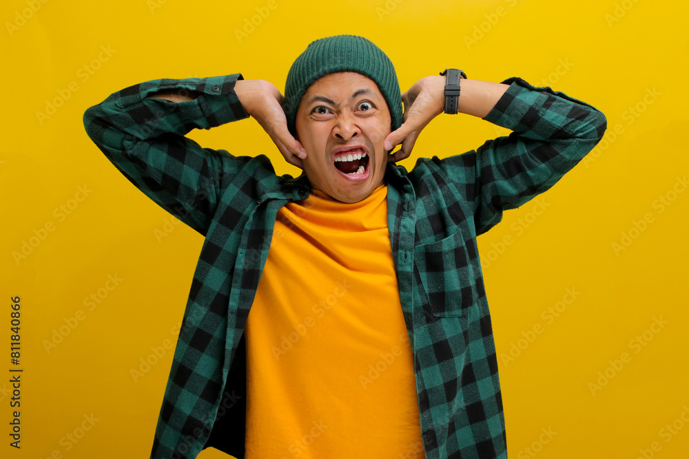 Thrilled and excited young Asian man, dressed in a beanie hat and casual shirt, grabs his head in disbelief, expressing both surprise and joy while standing against a yellow background Thrilled and excited young Asian man, dressed in a beanie hat and casual shirt, grabs his head in disbelief, expressing both surprise and joy while standing against a yellow background
