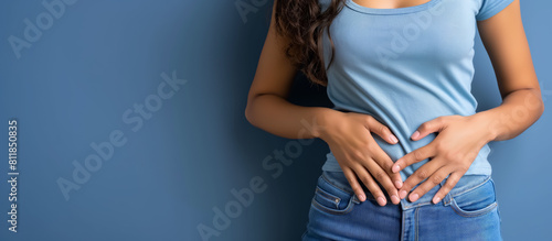 Woman holding her stomach with both hands, expressing discomfort or pain, set against a plain blue background, emphasizing her troubled expression.