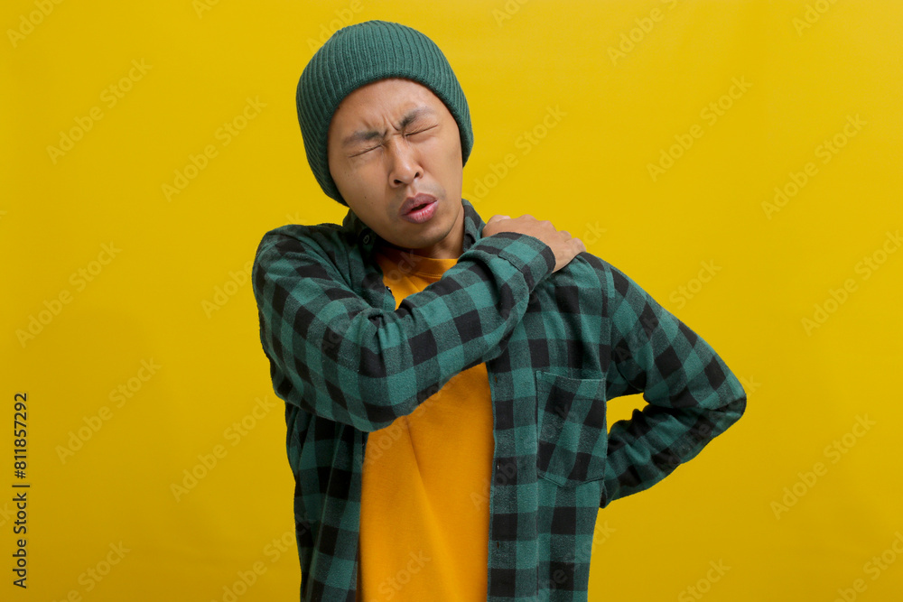 Asian man, dressed in a beanie hat and casual outfit, is suffering from neck and shoulder pain, isolated on a yellow background. Illustrating healthcare and problem concepts Asian man, dressed in a beanie hat and casual outfit, is suffering from neck and shoulder pain, isolated on a yellow background. Illustrating healthcare and problem concepts