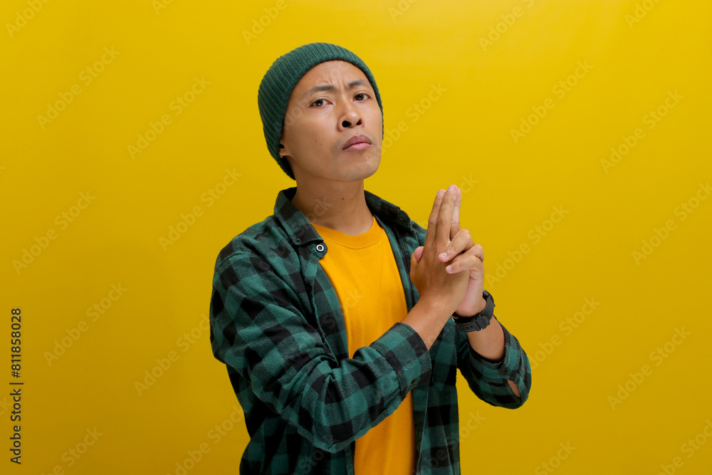 Young Asian man, dressed in a beanie hat and casual shirt, makes a finger gun gesture, mimicking shooting actions, with an angry expression, standing against a yellow background Young Asian man, dressed in a beanie hat and casual shirt, makes a finger gun gesture, mimicking shooting actions, with an angry expression, standing against a yellow background