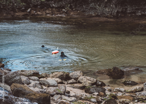 Two Scuba Divers Exploring Tranquil Waters Near Rocky Shoreline During Daylight